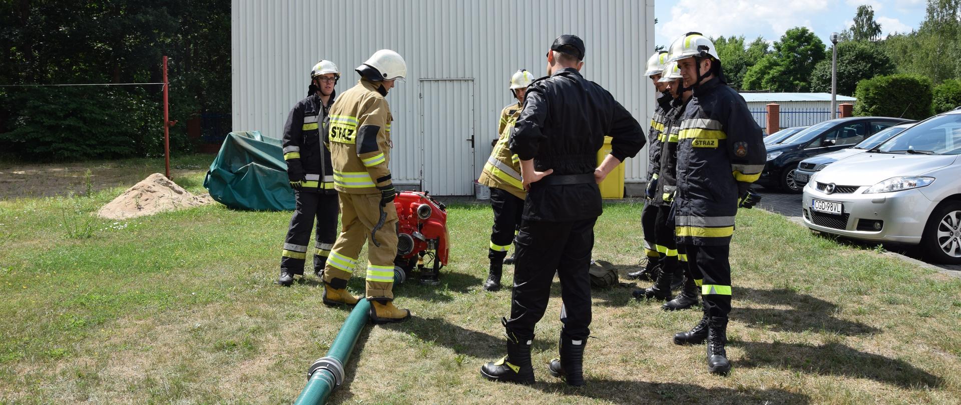 NA ZDJĘCIU WIDAĆ ODWRÓCONEGO TYŁEM STRAŻAKA W UMUNDUROWANIU DOWÓDCZO-SZTABOWYM, KTÓRY WRAZ ZE STRAŻAKAMI OSP UBRANYMI W UBRANIACH SPECJALNYCH STOI PRZY MOTOPOMPIE. W TLE WIDAĆ GARAŻ.
