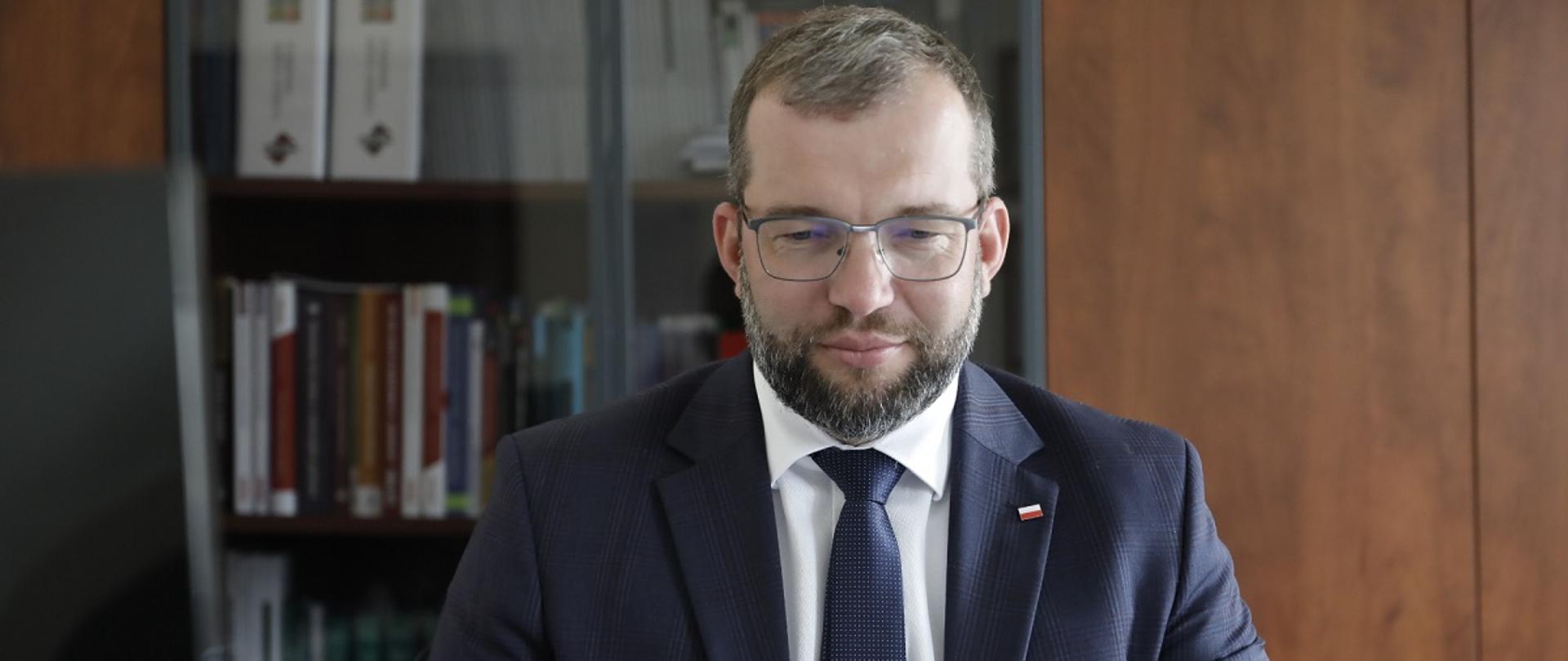 Minister funduszy i polityki regionalnej Grzegorz Puda na spotkaniu on-line Platformy Partnerstwa Publiczno-Prywatnego, minister siedzi przed laptopem, za jego plecami regał z książkami