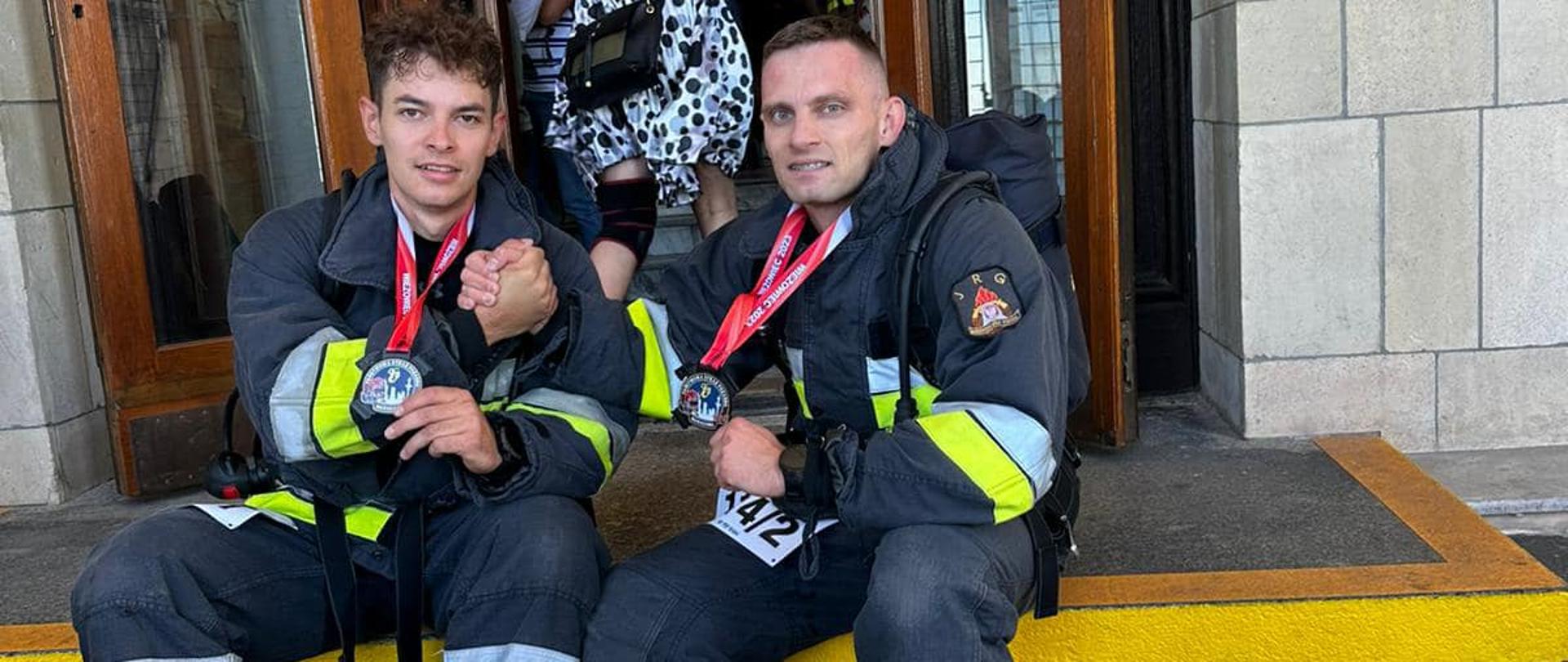 Dwóch strażaków po zakończeniu biegu po schodach siedzi z medalami na szyjach