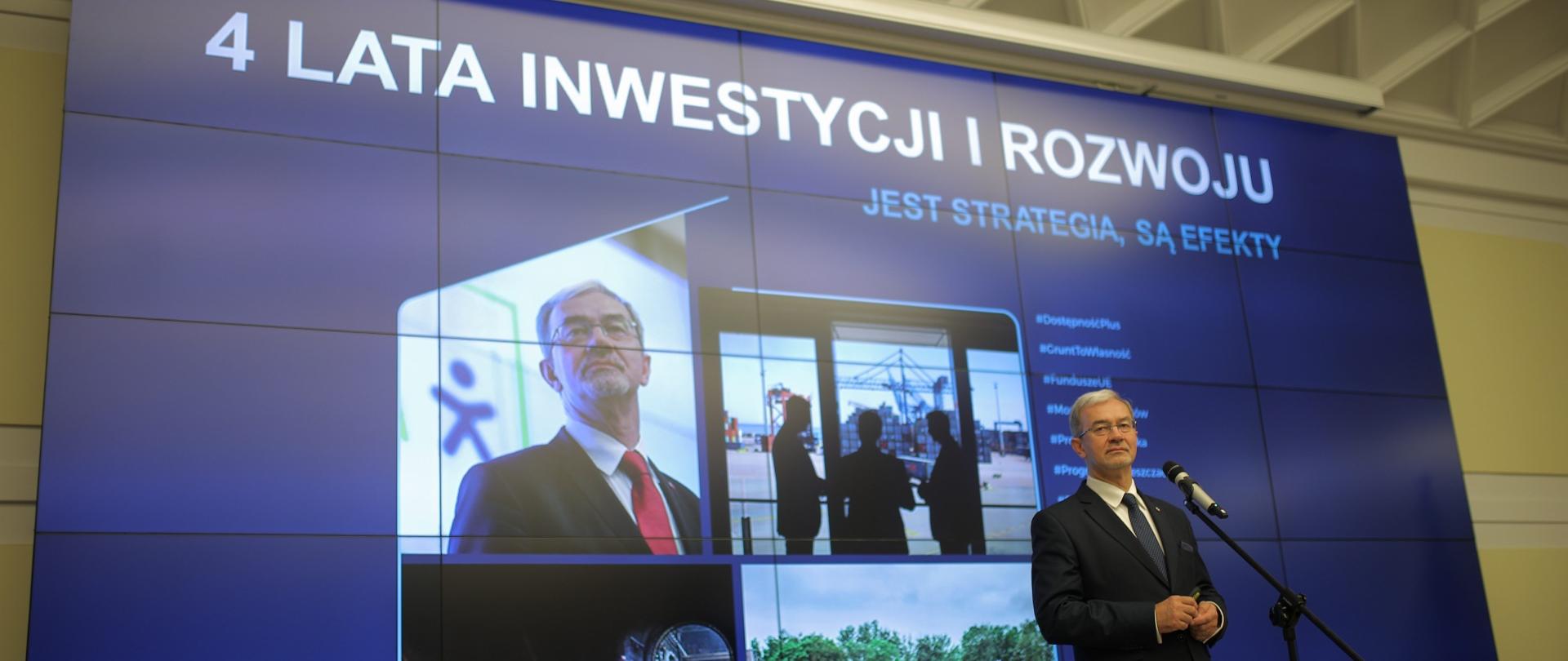 Minister Jerzy Kwieciński na scenie przy mikrofonie podczas konferencji, za nim ekran z grafiką ze zdjęciami związanymi z pracami ministerstwa oraz tytułem "4 lata inwestycji i rozwoju, jet strategia, są efekty"