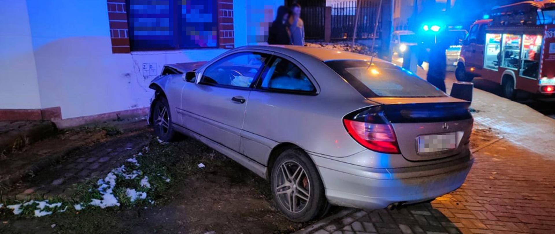 Widok z tyłu na rozbity samochód osobowy marki Mercedes. Na zdjęciu widoczny także radiowóz policji oraz samochód straży pożarnej. Widoczny niebieski zafarb od świateł błyskowych samochodów uprzywilejowanych.