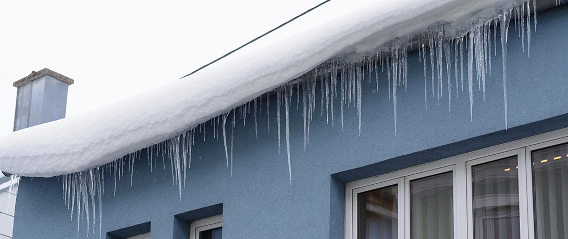 Zdjęcie przedstawia budynek, na dachu którego widać duże ilości śniegu oraz zwisające sople