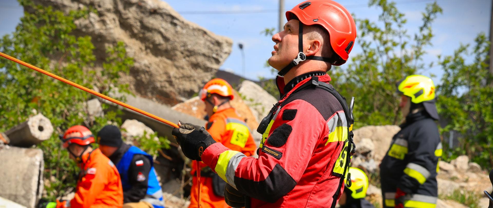 Ćwiczenia ratownicze pk. „POLIGON 2026” z udziałem sił i środków Centralnego Odwodu Operacyjnego Państwowej Straży Pożarnej z woj. podlaskiego - na zdj. strażak z grupy wysokościowej, trzymający linkę, ubrany w czerwony mundur, ma pas z zaczepami. W tle toczą się ćwiczenia.