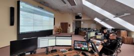 Sala stanowiska kierowania KG PSP, funkcjonariusze PSP siedzą przy biurkach i monitorach ekranowych, na ścianie przed nimi wisi duży ekran na którym wyświetlają się aplikacje z ćwiczeń