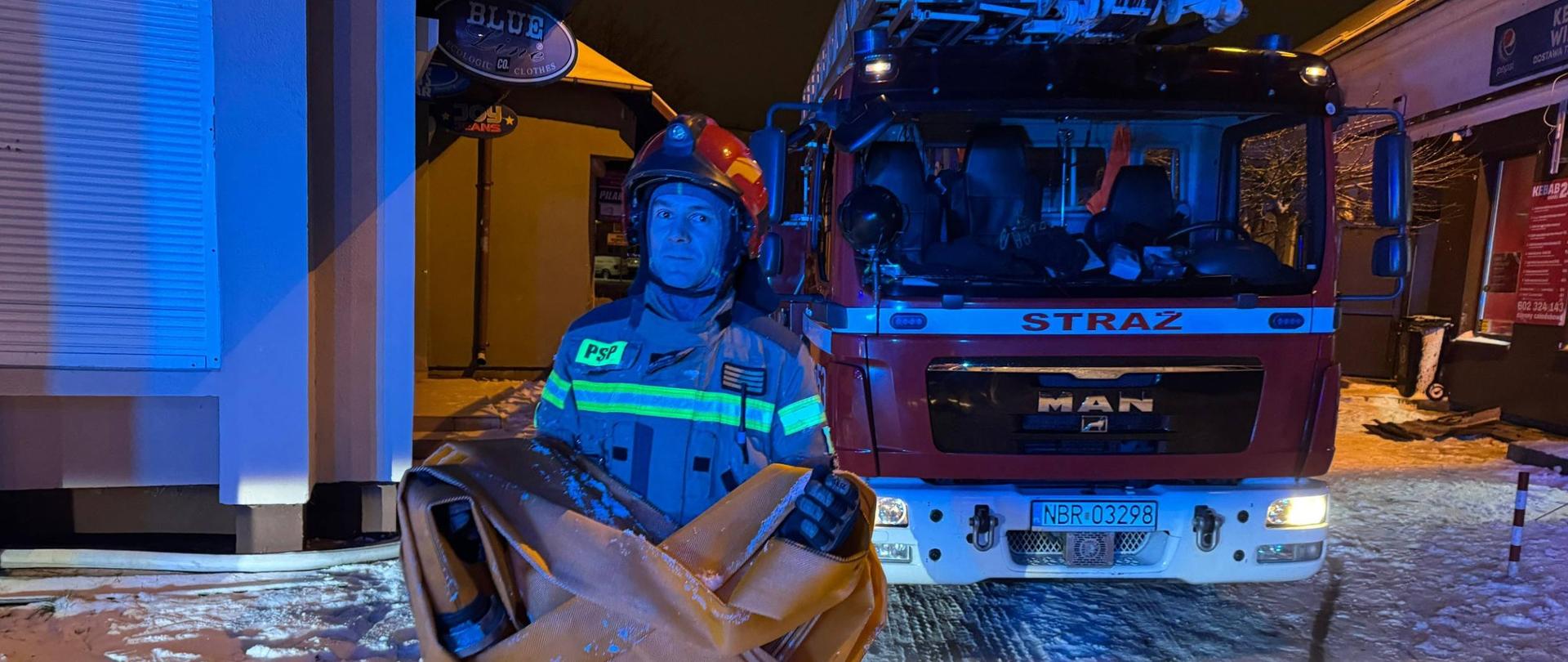 Strażak PSP niesie zwinięty wąż tłoczny na tle samochodu z drabiną mechaniczną SD; akcja ratowniczo-gaśnicza w zimowej scenerii nocą.