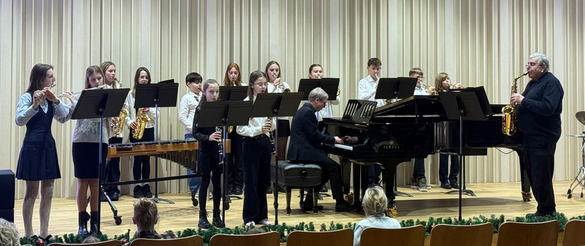 Na scenie sali koncertowej występuje młodzieżowa orkiestra szkolna. Uczniowie stoją i siedzą półkolem, grając na instrumentach dętych drewnianych i blaszanych oraz instrumentach perkusyjnych. W centrum widoczny jest fortepian koncertowy, przy którym siedzi akompaniator. Po prawej stronie dyrygent prowadzi orkiestrę. Muzycy ubrani są w stroje galowe. W tle jasna ściana akustyczna, na pierwszym planie fragment widowni.