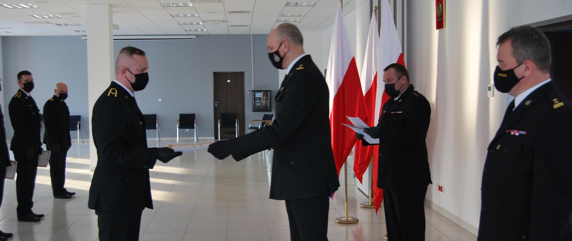 Wręczenie świadectw kursu zawodowego. Zastępca kujawsko-pomorskiego komendanta wojewodzkiego st. bryg. Slawomir Herbowski wręcza strażakom świadectwa ukończenia kursu zawodowego - pierwszego etapu szkolenia. . Wszyscy strażacy ubrani są w umundurowanie wyjściowe. Towarzyszą im flagi w barwie narodowej.