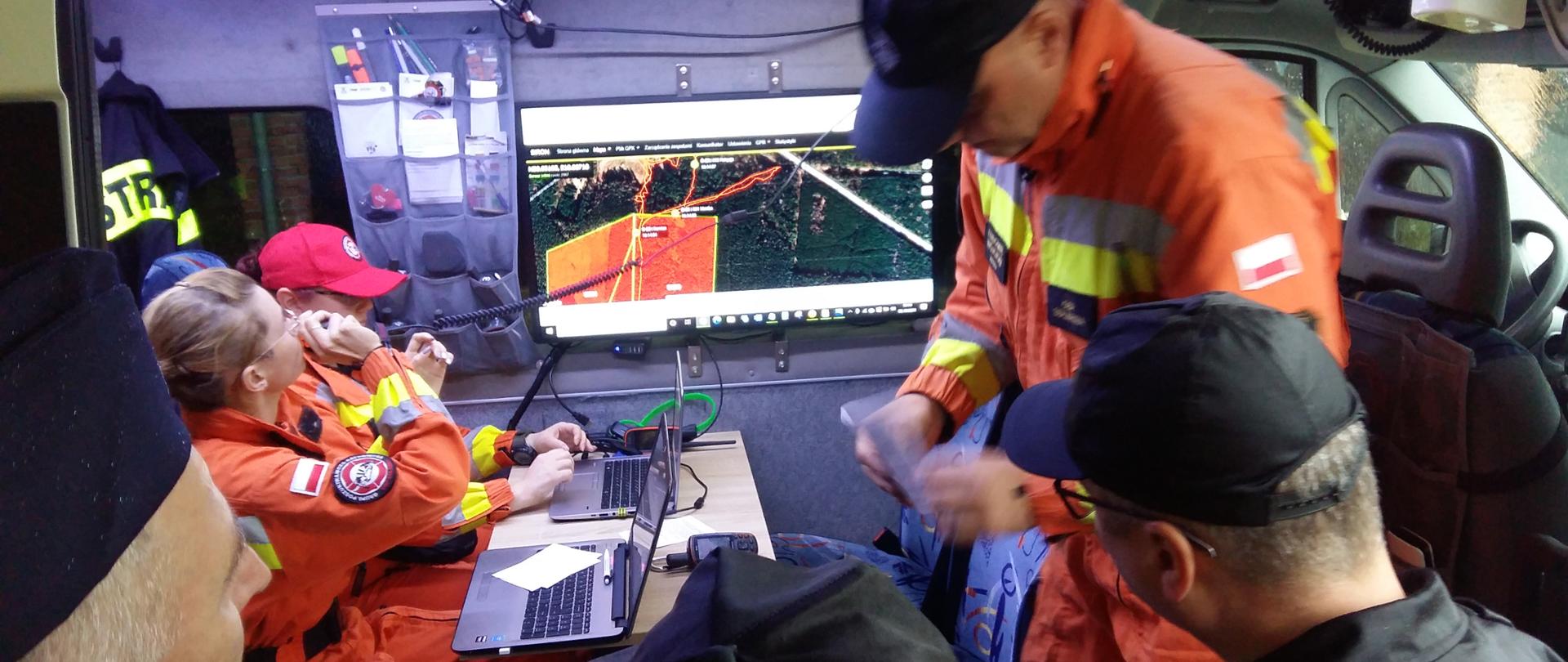 Strażacy podczas rozmowy na tle monitora z wyświetlonym terenem leśnym.