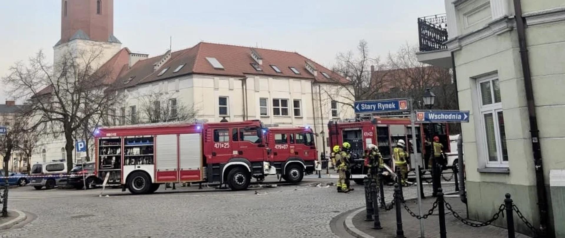 Wozy straży pożarnej ustawione na rynku miejskim, strażacy prowadzą działania przy budynku użyteczności publicznej. W tle widoczna wieża ratusza z zegarem, a teren akcji zabezpieczony jest przez służby ratownicze.