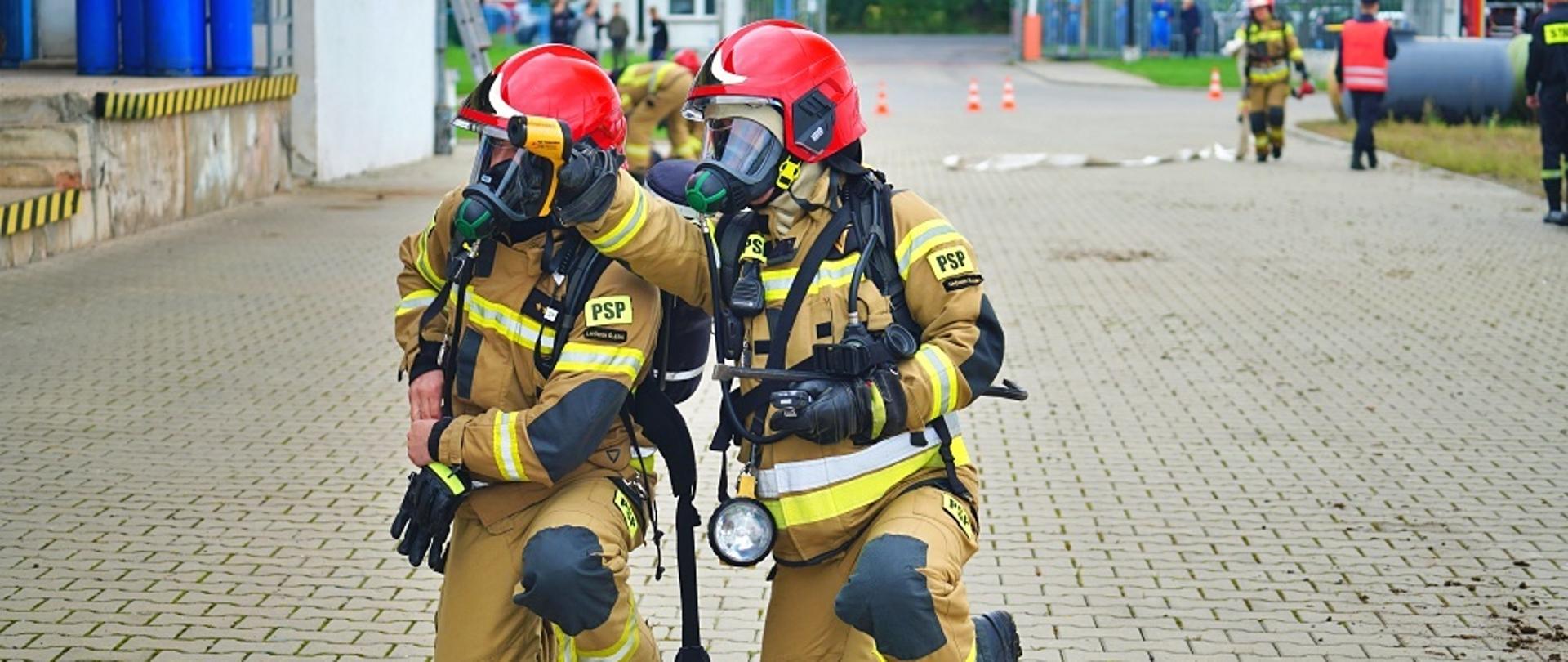 Dwóch strażaków prowadzi działania rozpoznawcze. W tle widoczni są inni strażacy biorący udział w ćwiczeniach.