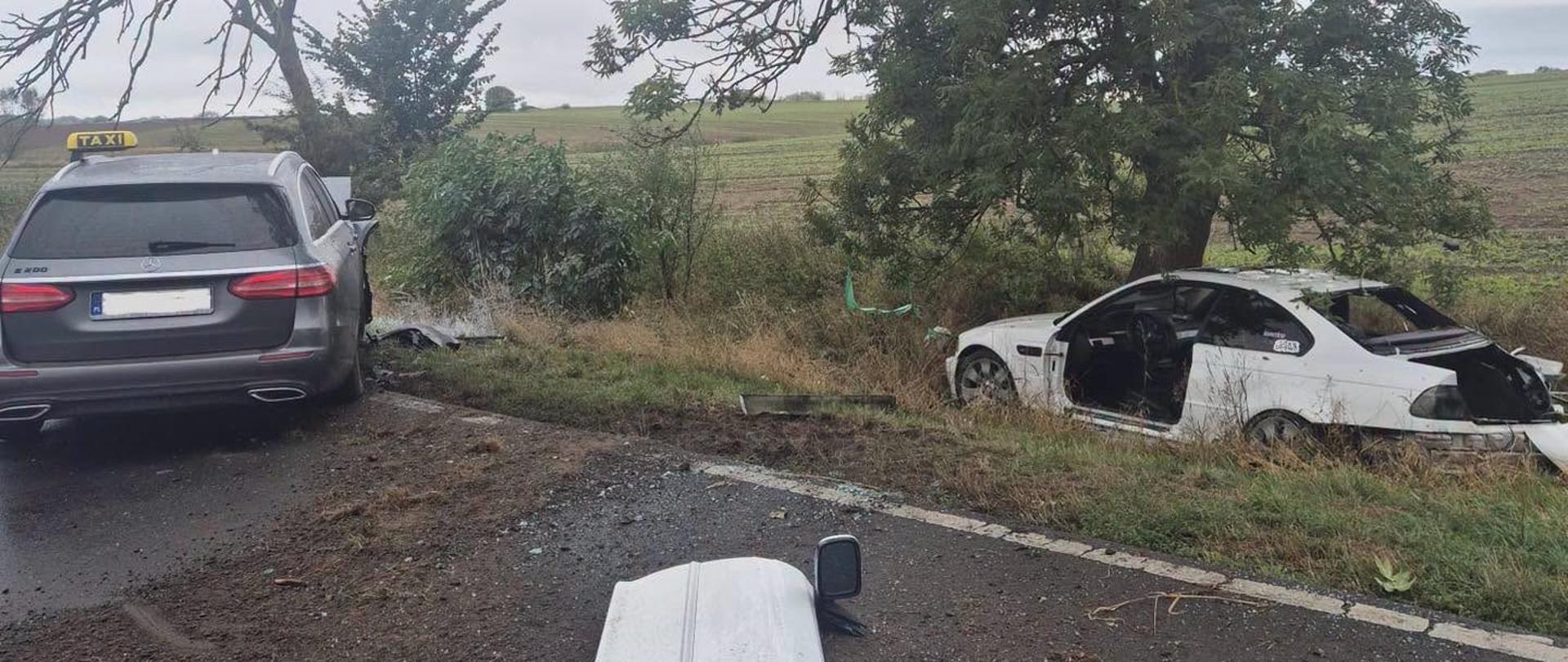 Miejsce wypadku drogowego. Uszkodzony biały samochód znajduje się poza jezdnią pod drzewem, a na drodze stoi samochód z oznaczeniem TAXI. Na jezdni i poboczu widoczne są elementy uszkodzonych pojazdów.