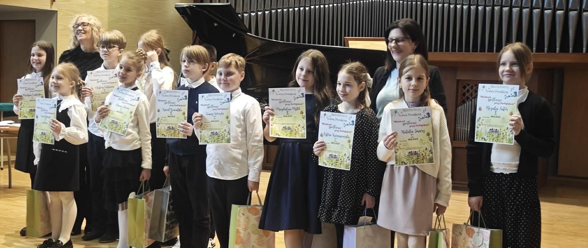 Zdjęcie przedstawia grupę dzieci i dorosłych na scenie sali koncertowej, po zakończeniu konkursu lub przesłuchania muzycznego. Dziesięcioro dzieci, ubranych odświętnie, stoi w rzędzie, trzymając w rękach dyplomy i torby z upominkami. Po lewej stronie widać dwie dorosłe kobiety, a w tle duży fortepian koncertowy i prospekt organowy. Scena ma drewnianą podłogę i boazerię na ścianach