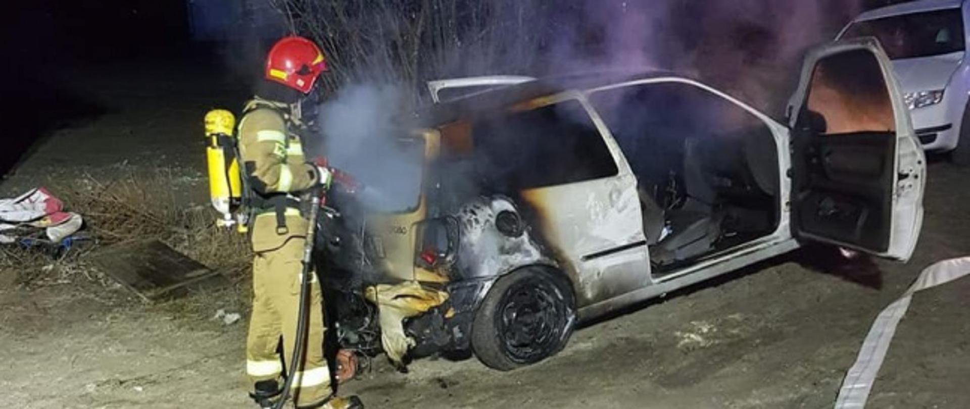 Zdjęcie przedstawia częściowo spalony samochód osobowy marki Volkswagen. Od strony bagażnika znajduje się strażak z linią szybkiego natarcia, który dogasza spalony pojazd. Strażak w ubraniu specjalnym oraz założonym na plecach aparatem ochrony dróg oddechowych. Z pojazdu wydobywa się lekkie zadymienie. W samochodzie otwarte są drzwi kierowcy oraz pasażera. Zdjęcie zrobione jest w porze nocnej.