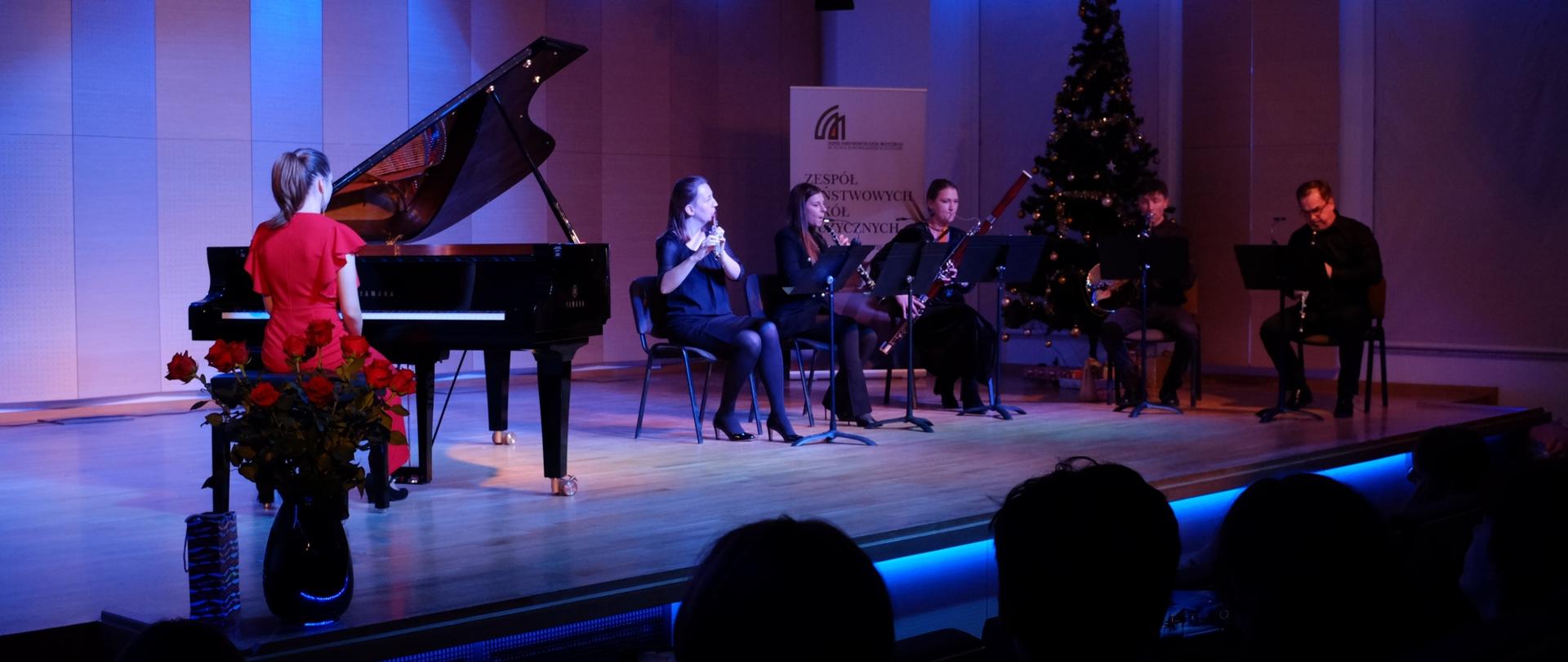 zdjęcie przedstawia kwintet dęty i solistkę przy fortepianie podczas wykonania utworu na scenie sali koncertowej, w tle baner szkoły