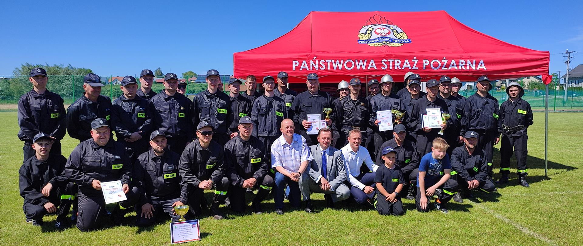 Zdjęcie przedstawia druhów OSP ustawionych w trzyszeregu. Ubrani są w ubranie koszarowe, pasy strażackie oraz hełmy lub czapki strażackie. Niektórzy z nich w dłoniach trzymają dyplomy i puchary. Po między nimi znajdują się 3 mężczyźni ubrani po cywilnemu. Za strażakami stoi czerwony namiot z napisem Państwowa Straż Pożarna. Wszystko dzieje się na boisku do piłki nożnej. 