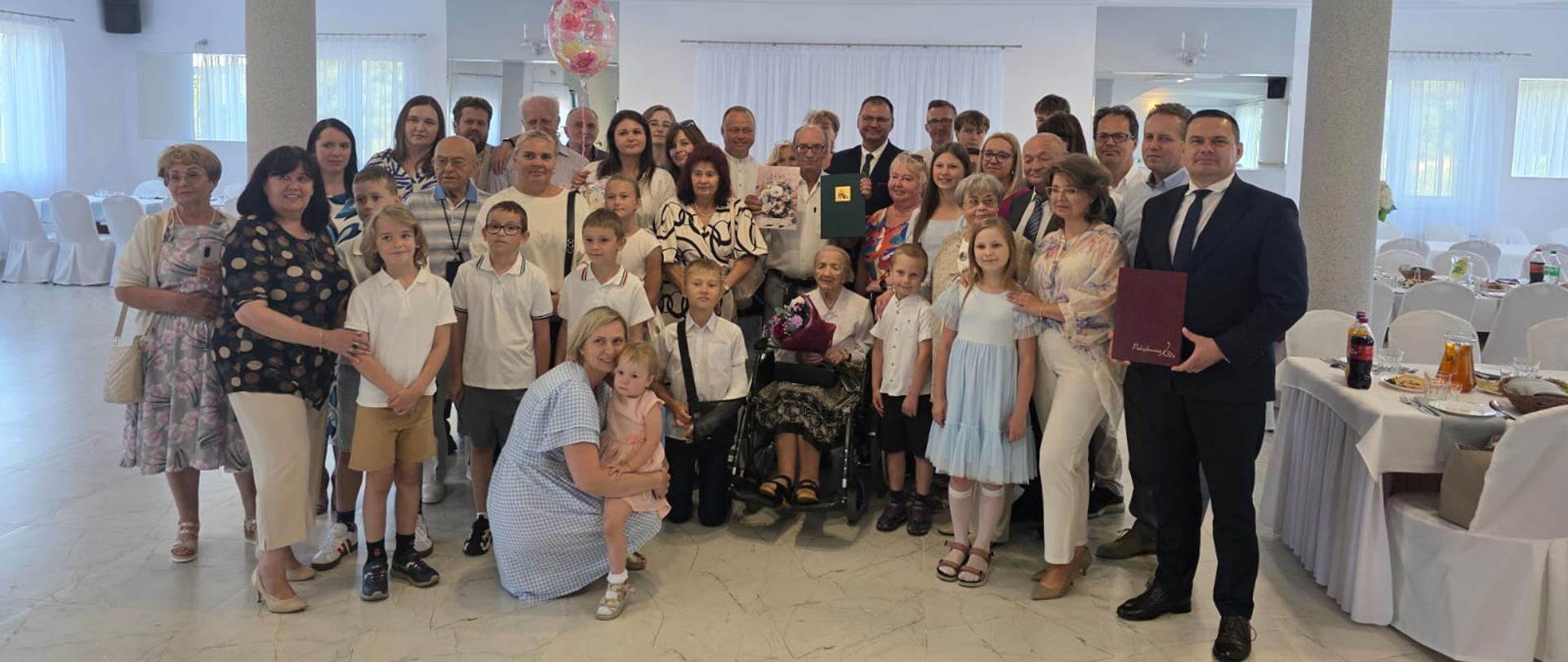 Grupa kilkudziesięciu osób, w tym kilkanaściorga dzieci pozuje do zdjęcia w udekorowanej na biało sali. Pośrodku grupy na wózku inwalidzkim siedzi elegancko ubrana starsza kobieta trzymająca w rękach bukiet kwiatów. Wszyscy uśmiechają się do zdjęcia.