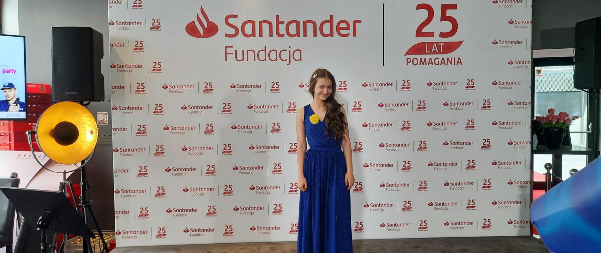 Antonina Waliszewska w niebieskiej sukience stoi na tle ścianki Fundacji Santander.