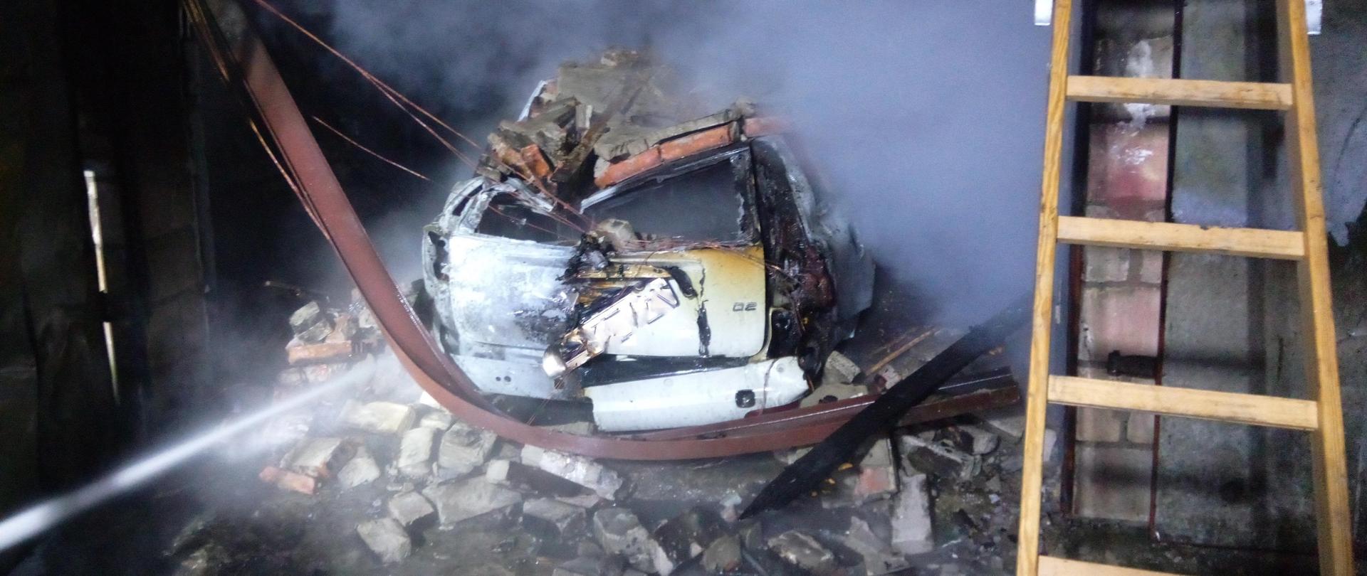 Wnętrze garażu, w środku spalony samochód, dach auta zgnieciony stalowo-betonową konstrukcją stropu.