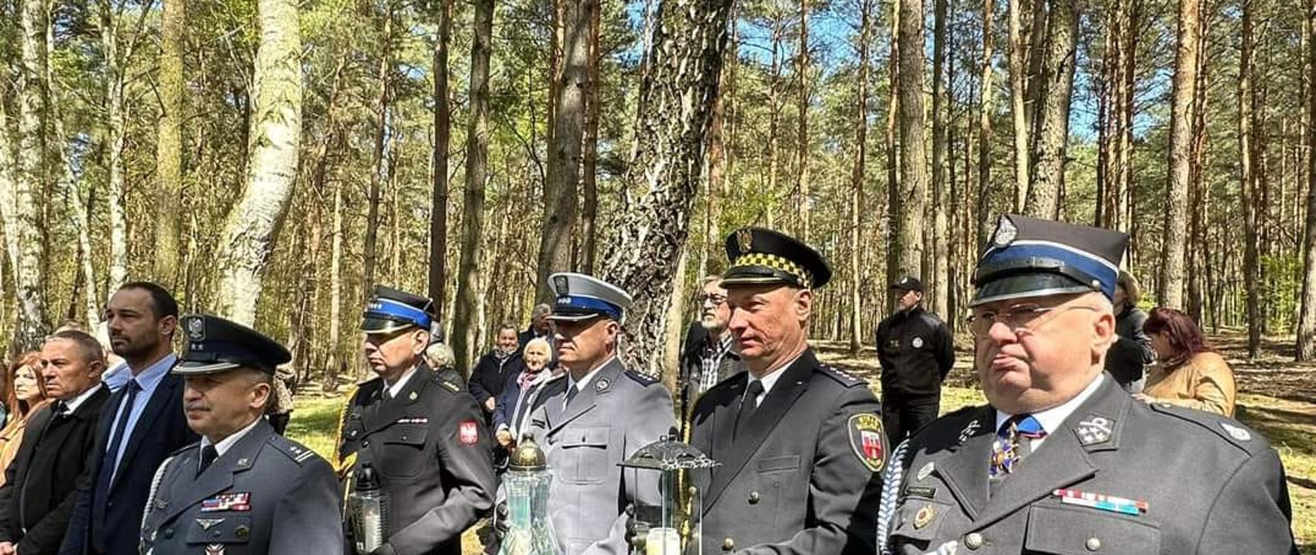 Uczestnicy uroczystości upamiętniających. Zdjęcie w ciągu dnia. Słonecznie. Widać las. Osoby umundurowane oraz w uraniach cywilnych.