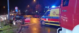 Samochód osobowy z uszkodzonym przodem stoi na poboczu. Obok na jezdni widoczni raotwnicy i pojazdy ratownicze.
