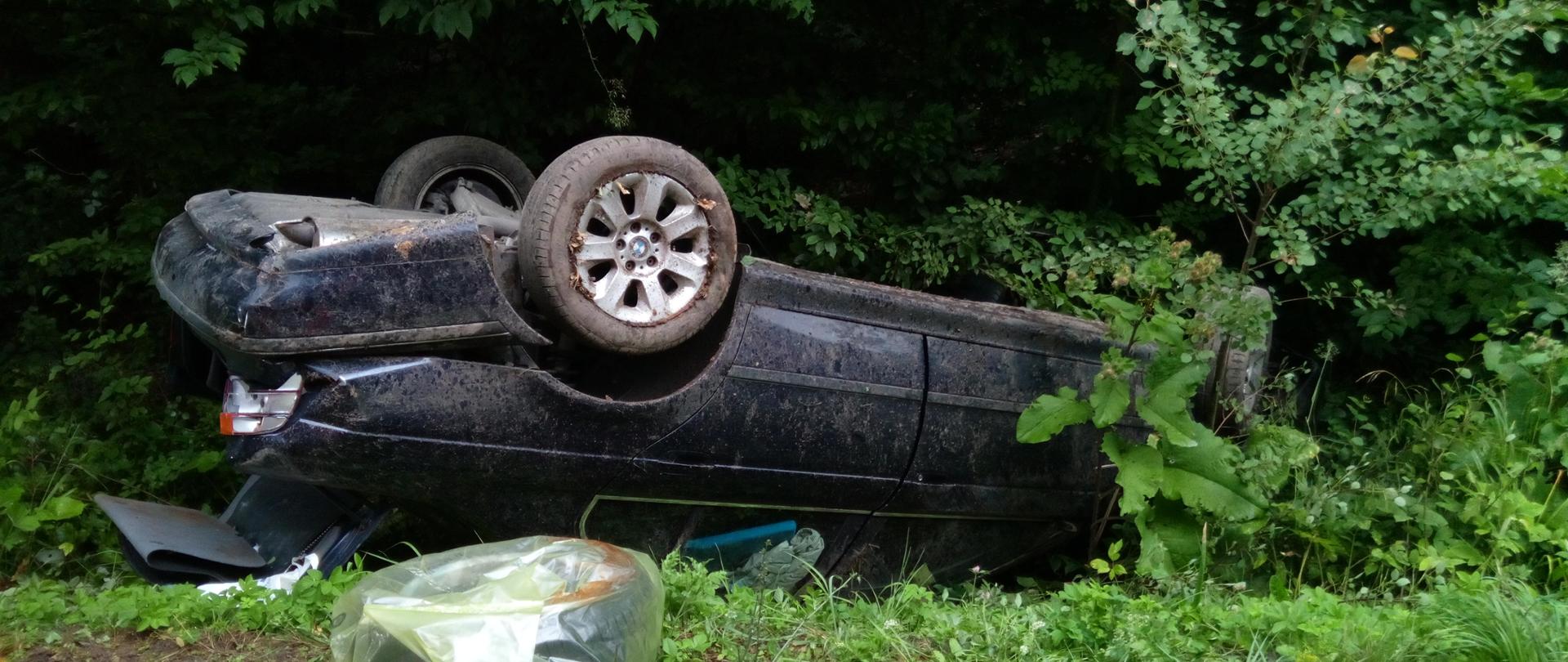 Zdjęcie przedstawia zniszczony samochód osobowy leżący na dachu w przydrożnym rowie.