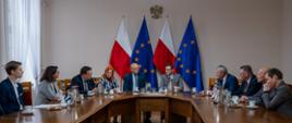 Kilkanaście osób siedzi wokół dużego, drewnianego stołu konferencyjnego w jasnej sali. Na ścianie w tle wiszą dwie flagi Polski, dwie flagi Unii Europejskiej oraz godło Polski. Na stole znajdują się dzbanki, filiżanki, szklanki i talerzyki z poczęstunkiem. 