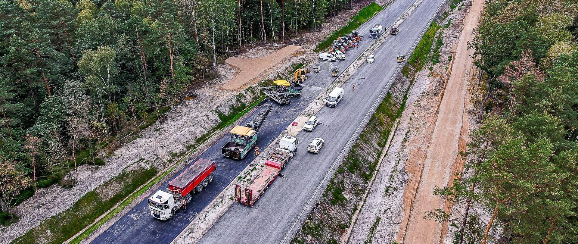 Zdjęcie przedstawia plac budowy drogi w lesie. Widoczna jest nowo budowana droga, na której pracują różne maszyny budowlane i pojazdy. Po lewej stronie drogi znajduje się las, a po prawej stronie widoczny jest pas ziemi przygotowany pod dalsze prace budowlane. Na drodze znajdują się ciężarówki, walce drogowe i inne maszyny budowlane. Zdjęcie zostało wykonane z lotu ptaka, co pozwala na szeroki widok na całą scenę.