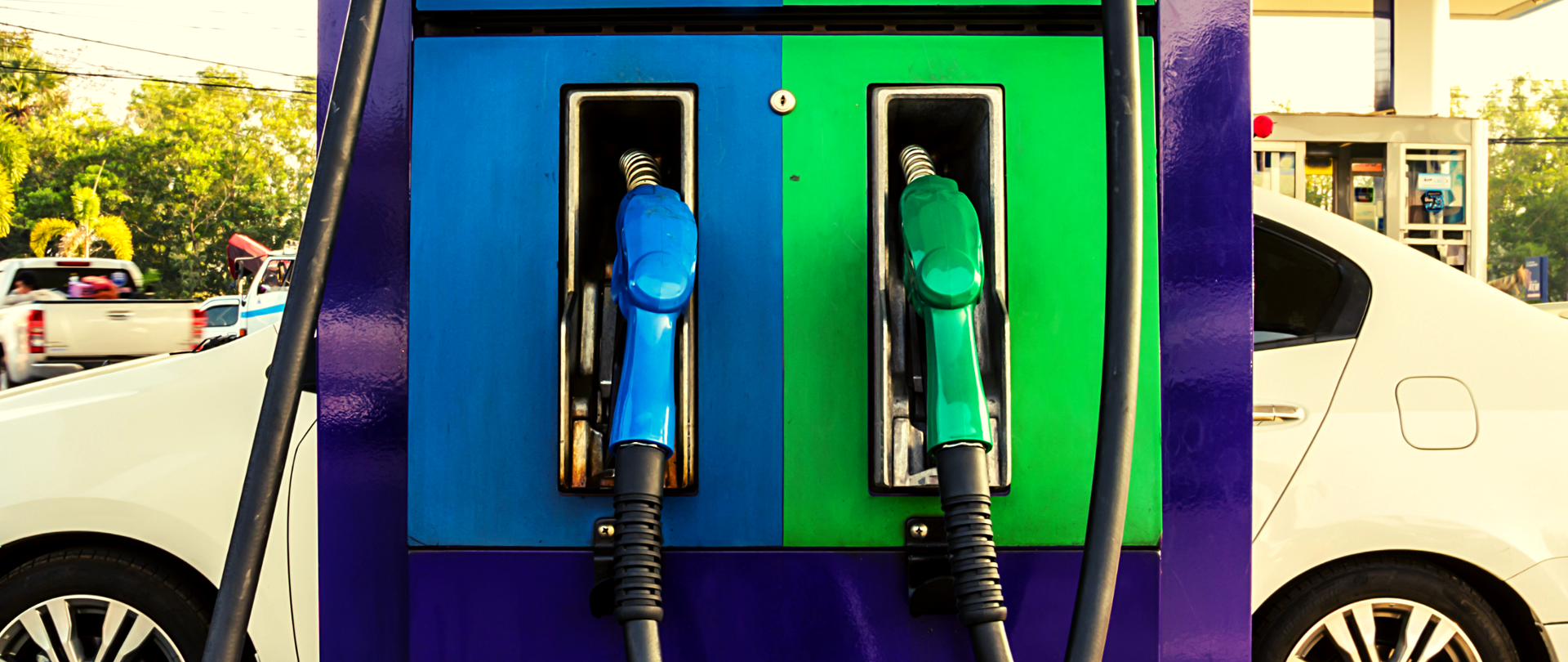 Na pierwszym planie niebiesko-zielony dystrybutor z paliwem, z tyłu stoi biały samochód