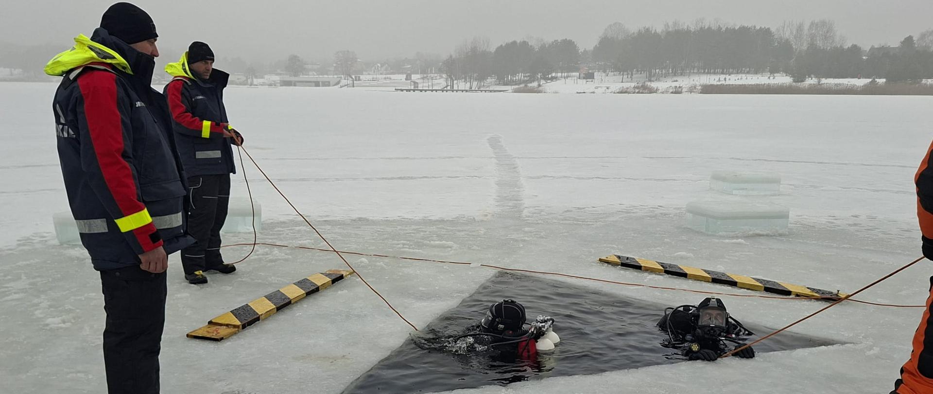Na zamarzniętym jeziorze w zimowej scenerii kilku ratowników w specjalistycznych strojach prowadzi ćwiczenia ratownictwa lodowego. Dwóch nurków w suchych skafandrach znajduje się w przeręblu z ciemną wodą, a pozostali ratownicy stoją na lodzie, asekurując ich linami. Wokół otworu leżą żółto-czarne belki zabezpieczające. W tle widać ośnieżony brzeg jeziora, drzewa i zabudowania, a nad całością panuje pochmurna, mroźna aura.