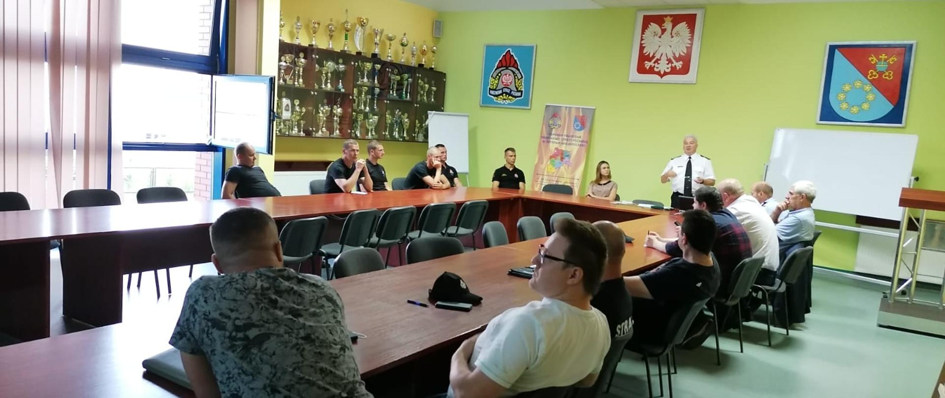 Funkcjonariusze i ochotnicy siedzący na sali narad w podkowie, Nad nimi stoi i przemawia komendant powiatowy.