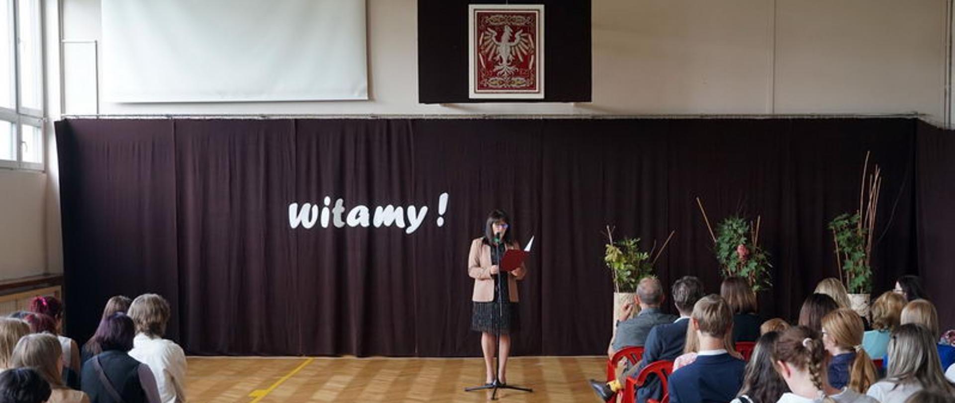 Zdjęcie przedstawia uczniów i dyrekcję PLSP w Kielcach na sali gimnastycznej. Pani Dyrektor przemawia przy mikrofonie. W tle widnieje napis "witamy" w kolorze białym na bordowym tle.