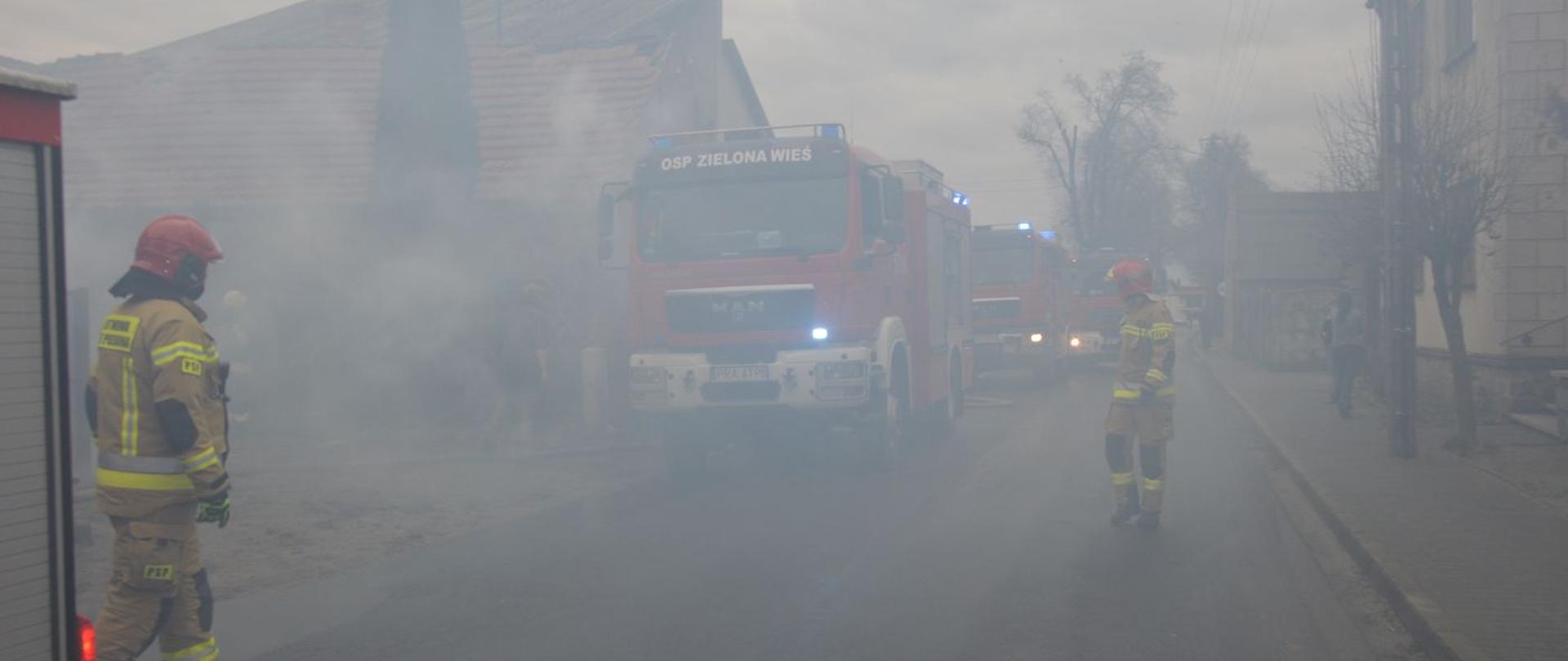Pożar budynku mieszkalnego w Wydawach. Zadymiona ulica, a na niej wozy strażackie z włączonymi sygnałami błyskowymi oraz dwóch strażaków w czerwonych hełmach. Po obu stronach drogi budynki.