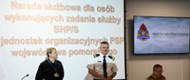 Pomorski komendant wojewódzki Państwowej Straży Pożarnej będący w białej koszuli oraz w spodniach z lampasami stoi, po lewej stoi funkcjonariusz w czarnym mundurze po prawej siedzi funkcjonariusz w mundurze w kolorze piaskowym. Za funkcjonariuszami na ekranie wyświetlany jest napis narada służbowa dla osób wykonujących zadania służby BHP/S jednostek organizacyjnych Państwowej Straży Pożarnej województwa pomorskiego. 