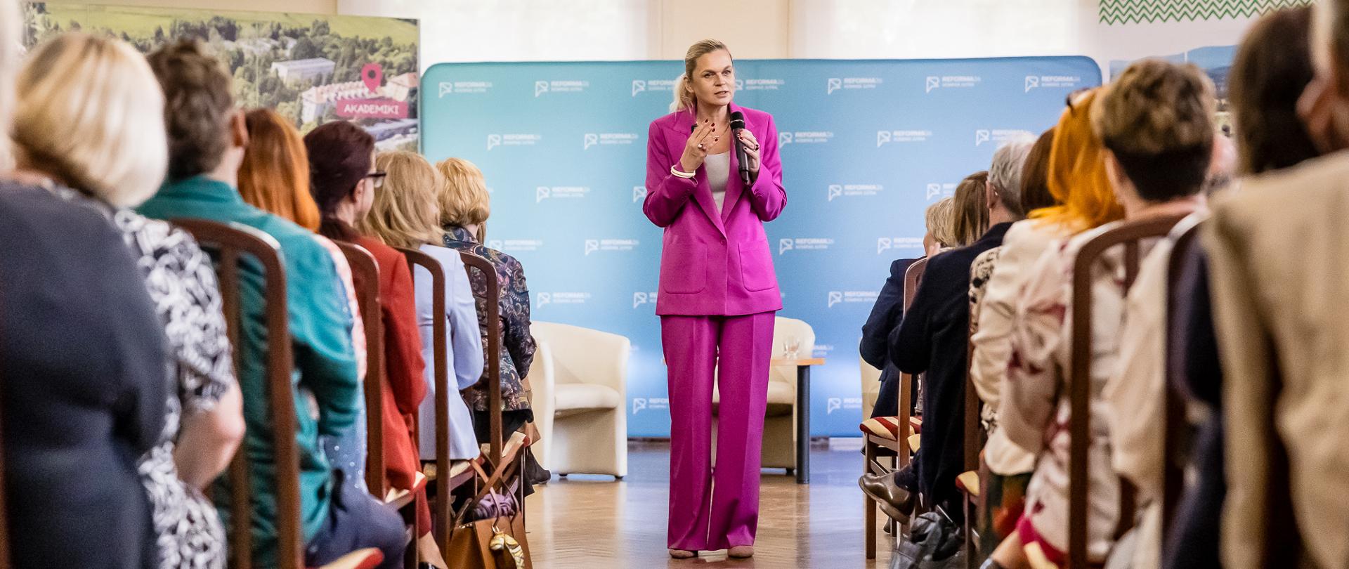Na zdjęciu Minister Edukacji Barbara Nowacka soi na środku sali i przemawia do nauczycieli podczas spotkania w Jeleniej Górze. Nauczyciele siedzą na krzesłach, w rzędach.