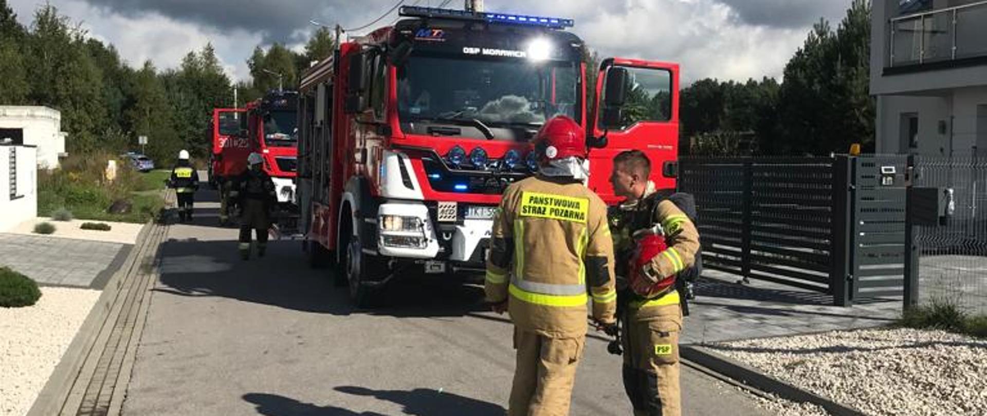 Zdjęcie przedstawia ulicę przy której doszło do pożaru domu. Po prawej dom, w którym doszło do pożaru. Na jezdni zaparowane dwa wozy strażackie, a przed nimi stoi dwóch strażaków.