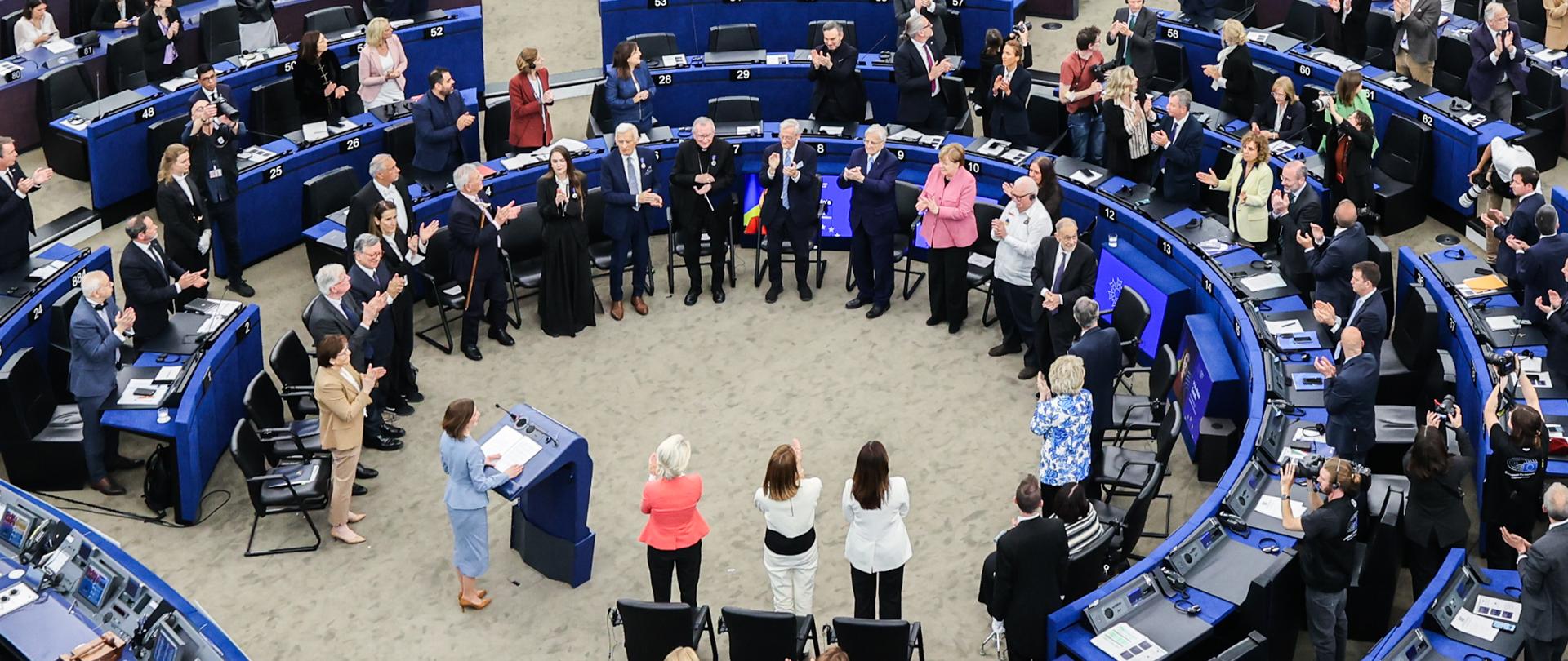 In the large European Parliament chamber, hundreds of participants stand beside semicircular rows of blue desks and applaud. In the center of the hall, a group of people is gathered, surrounded by parliament members and guests dressed in formal attire.