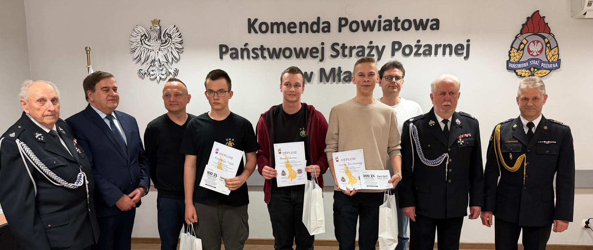 Zdjęcie grupowe w tej samej sali konferencyjnej. W pierwszym rzędzie stoi kilkanaścioro dzieci i młodzieży – uczestników turnieju. Każde z nich trzyma w rękach białą papierową torbę z upominkiem oraz pamiątkowy dyplom. Za nimi stoją dumni opiekunowie, strażacy oraz przedstawiciele komisji sędziowskiej. Po prawej stronie widoczny jest duży ekran multimedialny z napisem: „Ogólnopolski Turniej Wiedzy Pożarniczej – eliminacje powiatowe 2026”. Wszyscy patrzą w stronę aparatu, uśmiechając się.