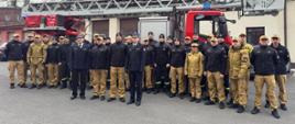 Na placu przed budynkiem komendy stoi liczna grupa strażaków w umundurowaniu służbowym. Funkcjonariusze ustawieni są w kilku rzędach i pozują do wspólnego zdjęcia. Bezpośrednio za nimi znajduje się czerwony samochód pożarniczy z drabiną mechaniczną. W tle widoczne są budynki jednostki oraz elementy infrastruktury przemysłowej