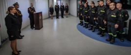 Zdjęcie zrobione w pomieszczeniu strażnicy. Po lewej stronie dwóch strażaków, Komendant Powiatowy stojący za mównicą, na wprost czterech strażaków ubrani w umundurowanie galowe. Po prawej stronie pododdział strażaków w umundurowaniu koszarowym.