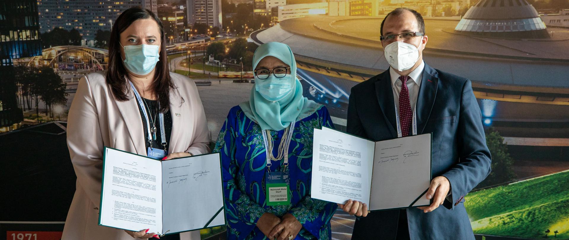 Na zdjęciu trzy osoby stoją na tle ścianki promocyjnej Katowic. Po lewej stronie wiceminister funduszy i polityki regionalnej Małgorzata Jarosińska-Jedynak prezentuje podpisaną właśnie umowę. Po prawej stronie tak samo robi dyrektor Instytutu Rozwoju Miast i Regionów Wojciech Jarczewski. W środku dyrektor wykonawcza UN Habitat Maimunah Mohd Sharif