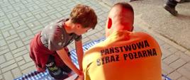 Strażak w piaskowym mundurze instruuje klęczącą osobę, jak prawidłowo wykonywać resuscytację krążeniowo-oddechową na fantomie. Widok z góry, zza pleców instruktora.
