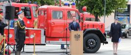 Wystąpienie przedstawiciela władz cywilnych przy mównicy z godłem państwowym, na tle zabytkowego samochodu pożarniczego. W tle widać również nowoczesne wozy bojowe oraz funkcjonariuszy w mundurach wyjściowych.
