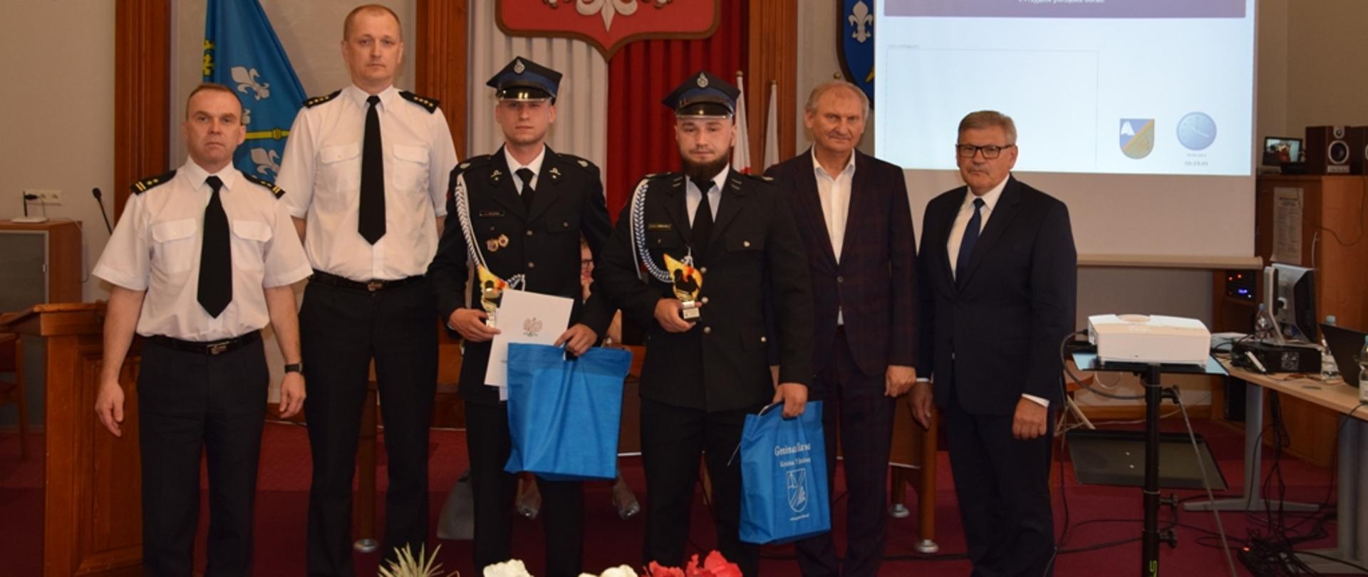 Komendant Powiatowy PSP w Iławie, ostródzki Zastępca Komendanta Powiatowego PSP i Wójt Gminy Iława wraz z wyróżnionymi strażakami OSP Rudzienice pozują do pamiątkowego zdjęcia. 