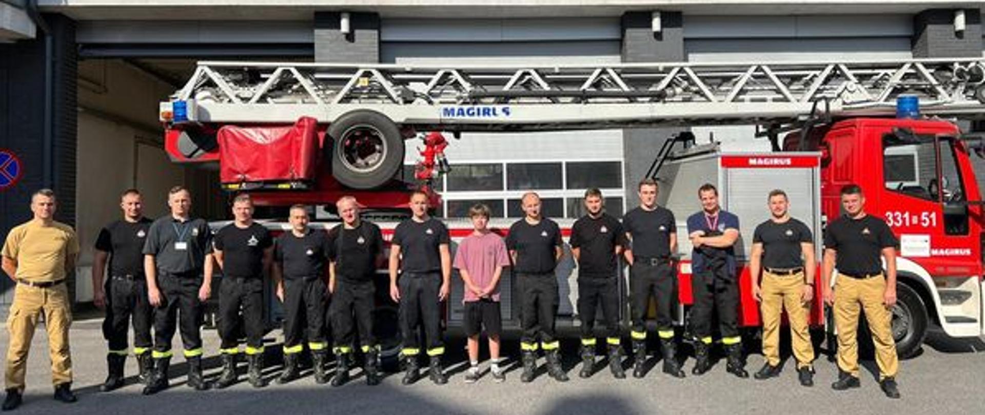 Zdjęcie wykonane przed budynkiem jednostki ratowniczo-gaśniczej numer 1 w Radomiu. Na zdjęciu widać strażaków oraz jednego z chłopców, który wylicytował dzień w komendzie miejskiej w ramach wielkiej orkiestry świątecznej pomocy. Za nimi w tle ustawiony jest pojazd specjalny drabina mechaniczna.