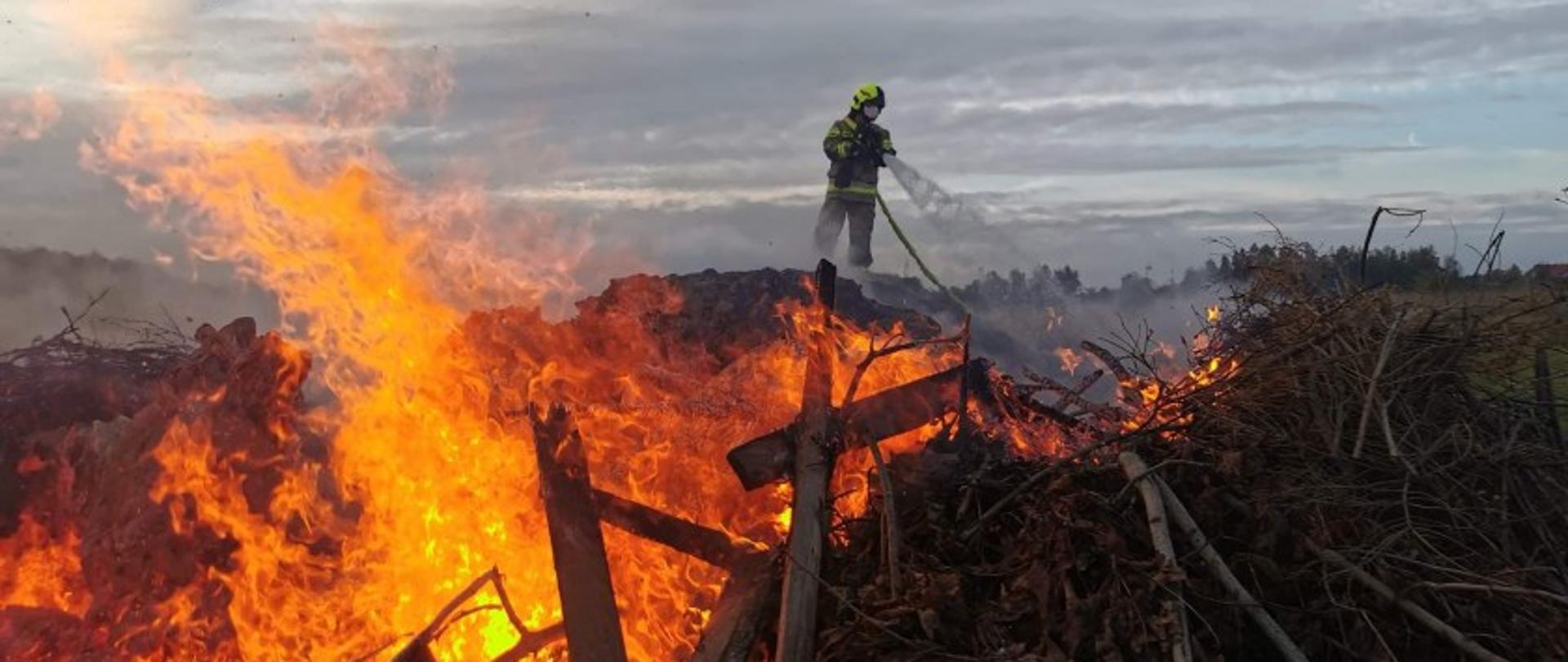 Paląca się sterta roślinności, na górze strażak gasi pożar.