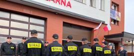 Przed budynkiem komendy stoją w dwóch rzędach strażacy. Na budynku komendy widoczny baner z napisem dbamy o twoje bezpieczeństwo 