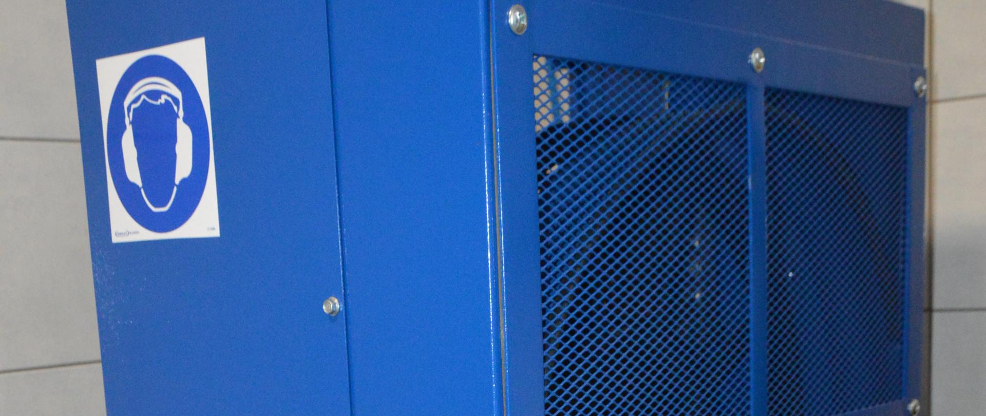 Zaplecze sprzętu ochrony układu oddechowego w Komendzie Powiatowej PSP w Rawiczu. Na planie głównym nowa sprężarka do ładowania butli. Urządzenie ma kształt prostopadłościanu. Jest koloru niebieskiego. Z lewej strony widoczny jest czarny wężyk. 