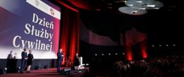 Widok sceny i części sali widowiskowej wraz z publicznością, na scenie osoby przemawiające na tle napisu "Dzień Służby Cywilnej"