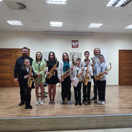 Ośmioro dzieci - cztery dziewczynki i czterech chłopców- oraz dwóch dorosłych stoi na scenie. Są to uczniowie klasy saksofonu, ich nauczyciel oraz akompaniator. Wszyscy uśmiechają się po udanym koncercie. Dzieci trzymają swoje saksofony. Dwie dziewczynki są ubrane w sukienki. Jedna założyła marynarkę, koszulę i spódnicę. Kolejna ubrała się w długi sweter i spodnie. Chłopcy również prezentują się bardzo elegancko. Wszyscy założyli koszule i ciemne spodnie. Jeden z nich dodatkowo przywdział muszkę. Nauczyciel jest ubrany w szary golf i spodnie, akompaniator w czarną koszule i spodnie. W tle na ścianie godło polskie fragment wizerunku Jana Sebastiana Bacha oraz grafiki partytury. 