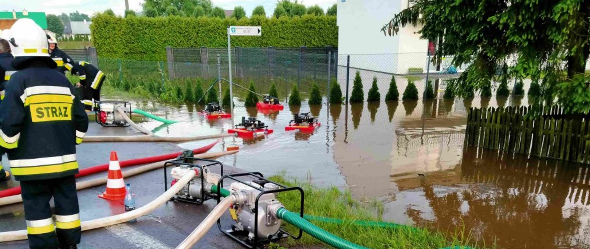 Na zdjęciu widoczna część drogi oraz fragmenty dwóch ogrodzonych posesji, które są podtopione. Woda wypompowywana jest przez osiem pomp: cztery pływające oraz czerty stojące na ziemi. Stojący na jezdni strażacy nadzorują pracę pomp.